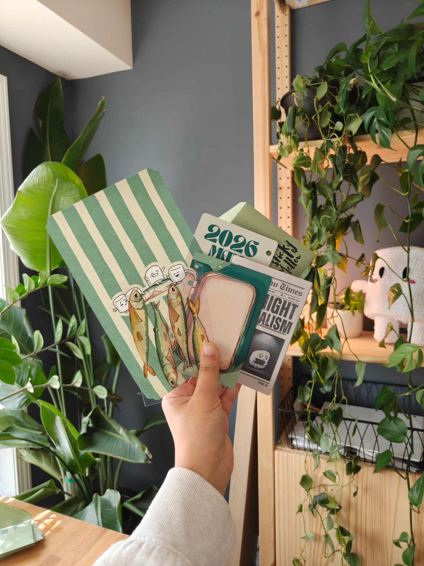 A hand holding a "sticky and mellow" collage of Aprils mail that consists of, a mini scenery 3D card along with its stickers themed marshmallow and sardine, a striped green beige cover and mini newspaper, and a green envelope, in a room with plants